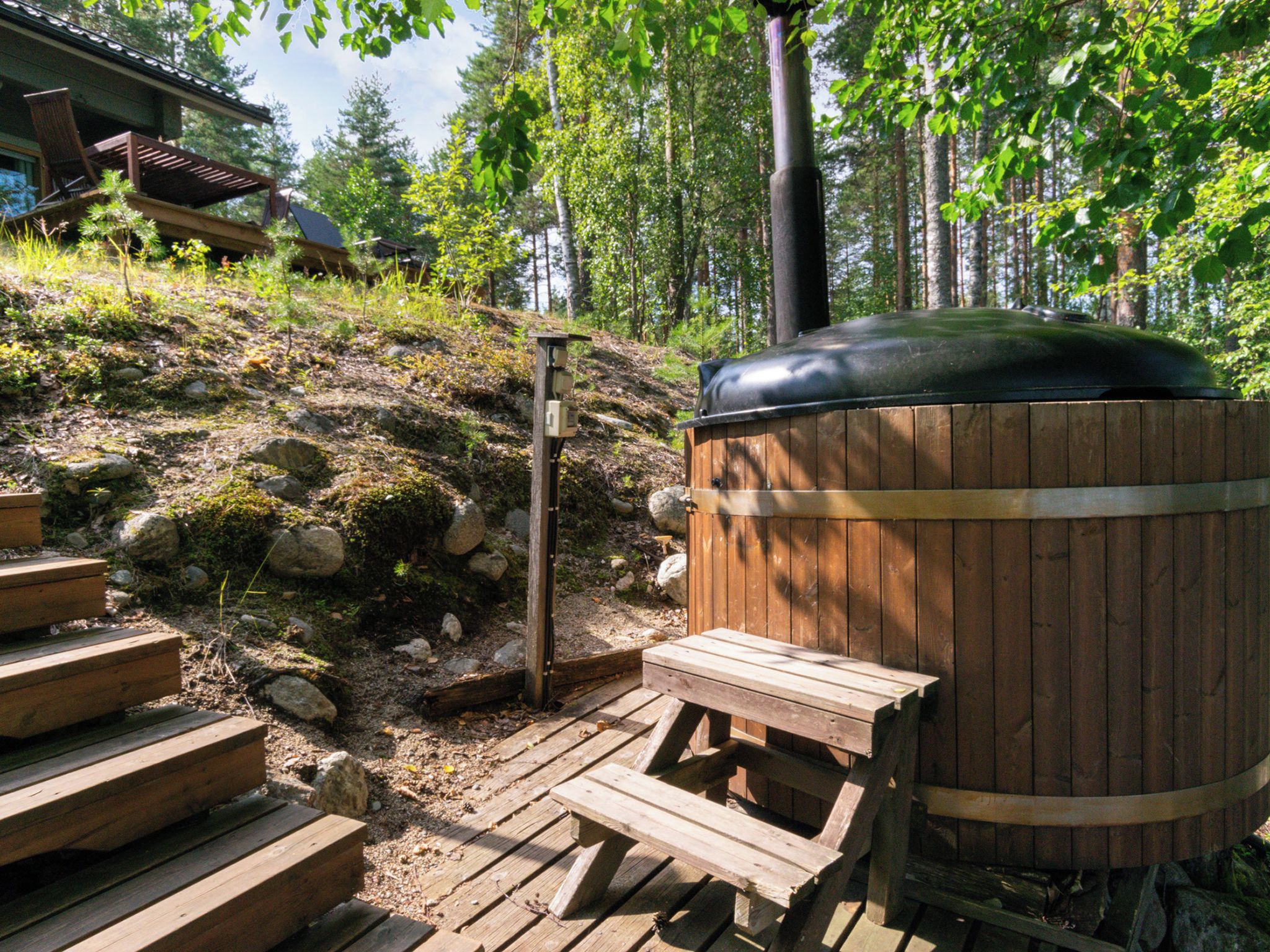 Una terraza de madera con un jacuzzi, escalones de madera y una ladera boscosa al fondo.