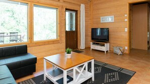 A wooden cabin interior with a sofa, a table with a plant, a TV on a stand, and large windows.