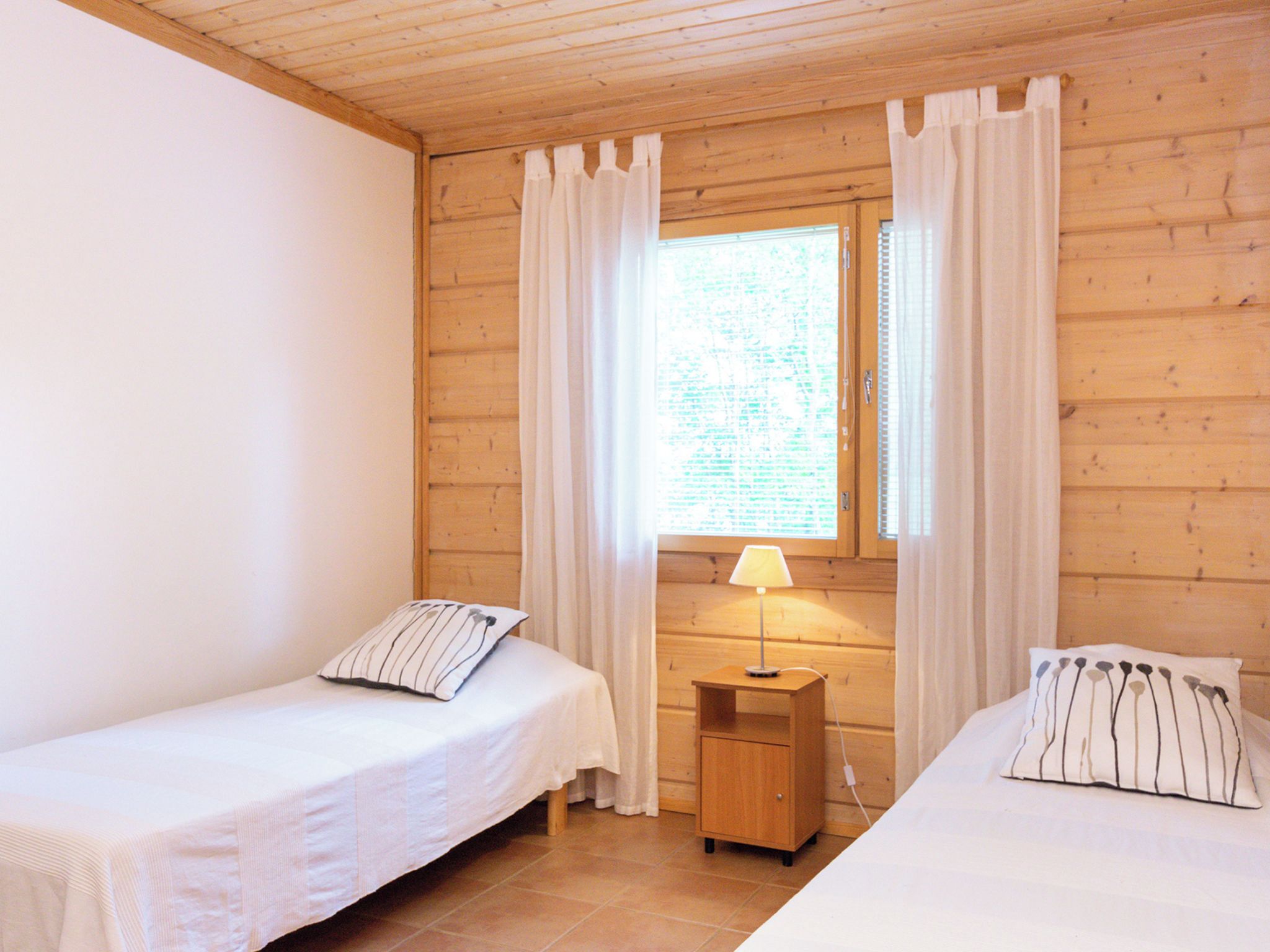 A room with two single beds, a nightstand with a lamp, and a window with curtains.