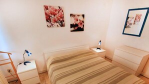 Een slaapkamer met een bed, een nachtkastje, een dressoir, een bureau en een bureaulamp. Twee ingelijste foto’s hangen aan de muur.