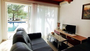 Un salon avec un canapé en cuir noir, une table basse en verre, un téléviseur posé sur un meuble en bois et une vue sur une piscine, visible à travers une porte vitrée.
