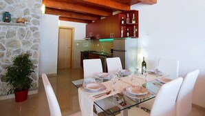 Un espace repas moderne avec une table en verre pour quatre personnes, des chaises blanches, un mur en pierre, des poutres en bois et une cuisine en arrière-plan.