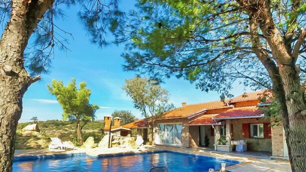 Un espace aménagé au bord de la piscine avec une table et des chaises, un bâtiment en pierre avec un toit en tuiles et un ciel bleu dégagé.