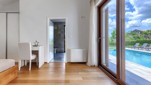 A modern hotel room with a wooden floor, a desk with a chair, a bed, a large sliding glass door leading to a pool area with lounge chairs.