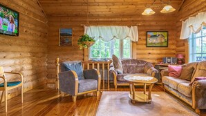 A cozy cabin interior with wooden walls, a wooden floor, a sofa, two armchairs, a small table, a hanging plant, and a television.