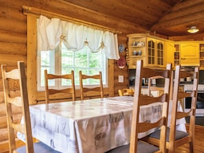 Een houten blokhut van binnen met een eettafel, stoelen, een keukenhoek en een raam met gordijnen.