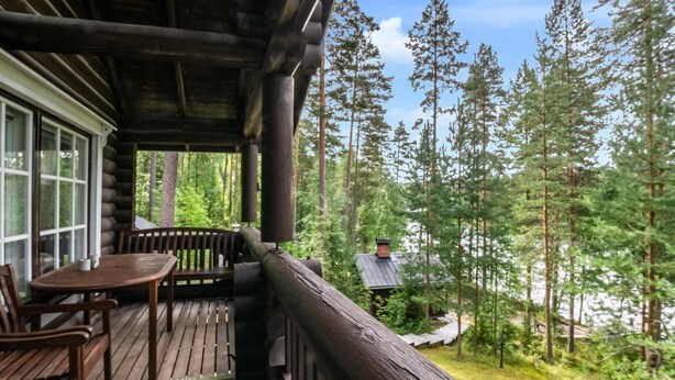 A wooden deck with a table and chairs, overlooking a forested area with tall trees and a cabin in the background.