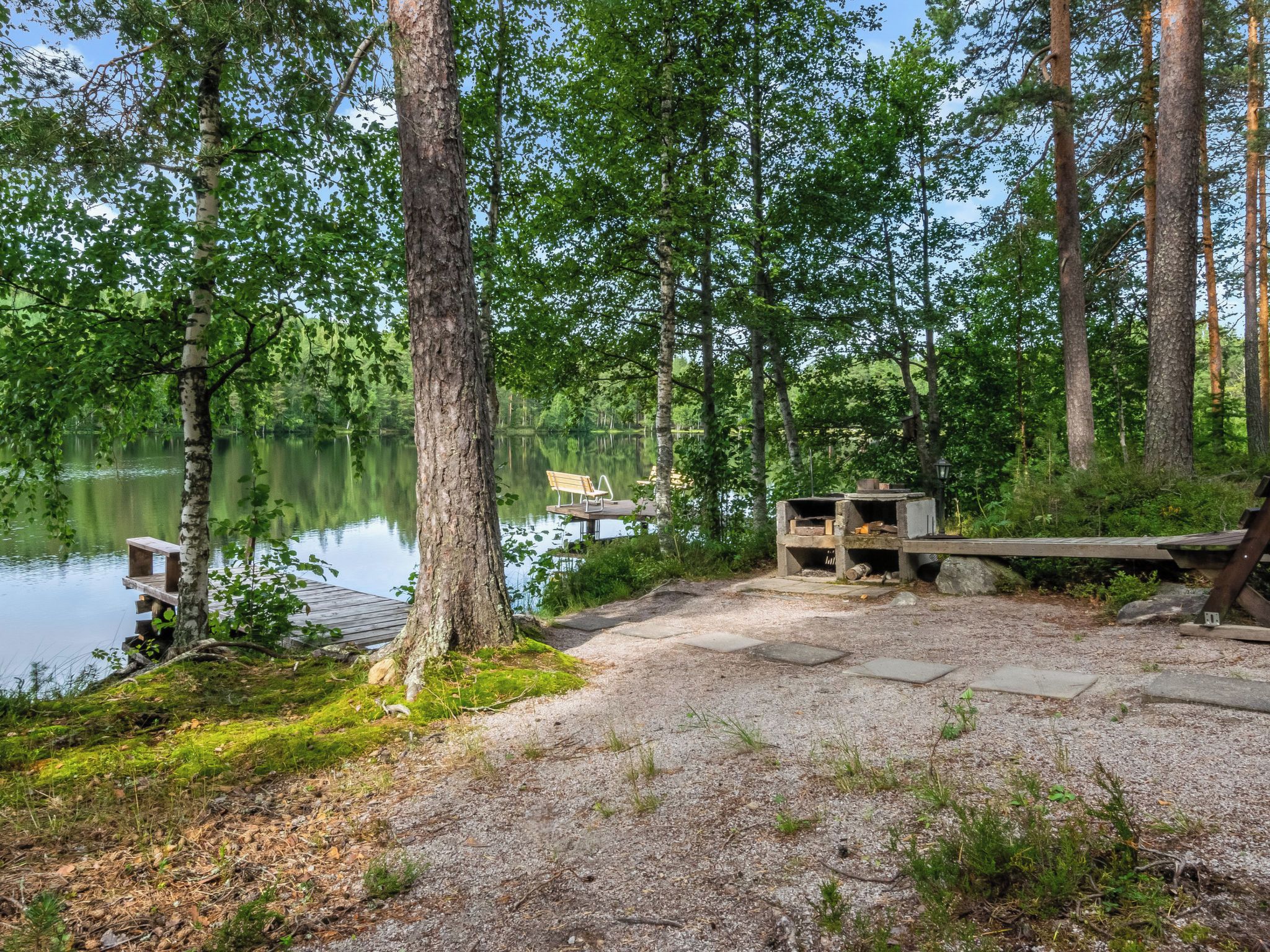 Rauhallinen leirintäalue järven rannalla, jossa on puinen penkki, piknikpöytä ja pieni suoja.