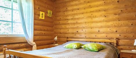 Un dormitorio de cabaña de madera con una cama, dos almohadas, una mesita de noche, una lámpara y una ventana con cortinas.