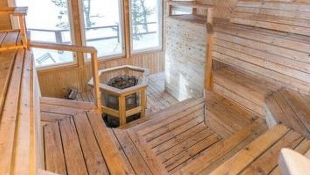 A wooden cabin interior with a central fireplace, wooden benches, and a window overlooking a snowy landscape. - Hästöskata b by Interhome (Kruunupyy)
