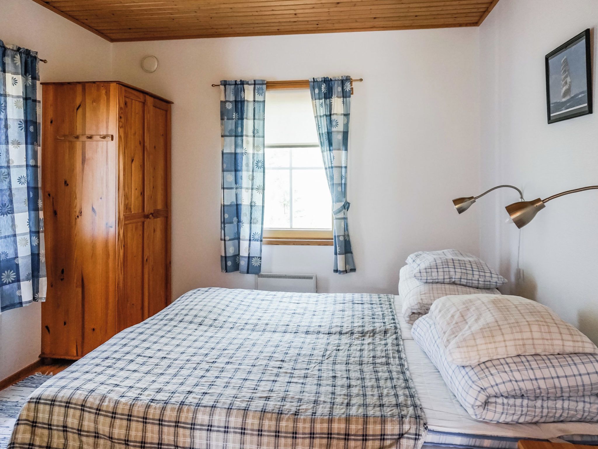 A bedroom with a wooden wardrobe, a window with curtains, a bed with a plaid bedspread, and a bedside lamp.