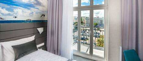 Une chambre d’hôtel avec un lit, une vue sur la ville grâce à de grandes fenêtres, et une fresque représentant le ciel et la mer sur un des murs.