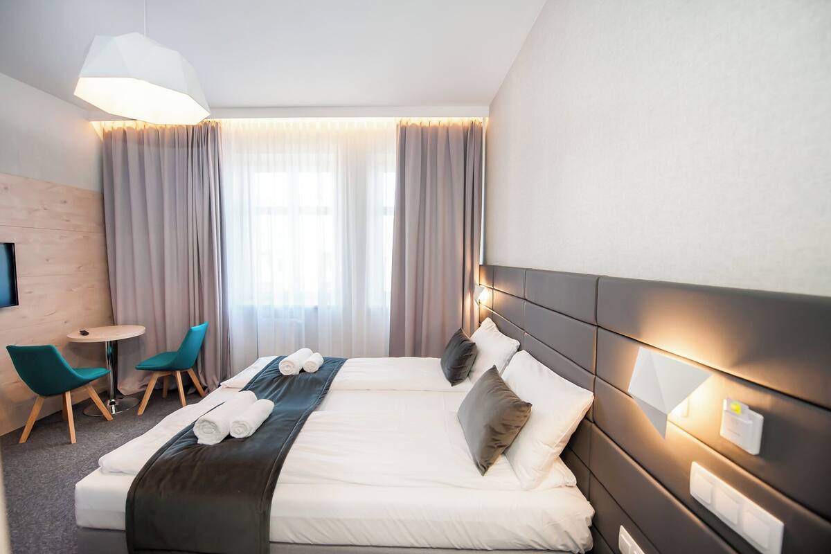 A modern hotel room with a large bed, a small round table, two teal chairs, and a TV mounted on the wall.