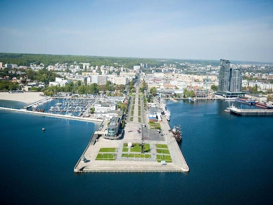 En vy från ovan över en småbåtshamn med båtar, en central strandpromenad och omgivande byggnader.
