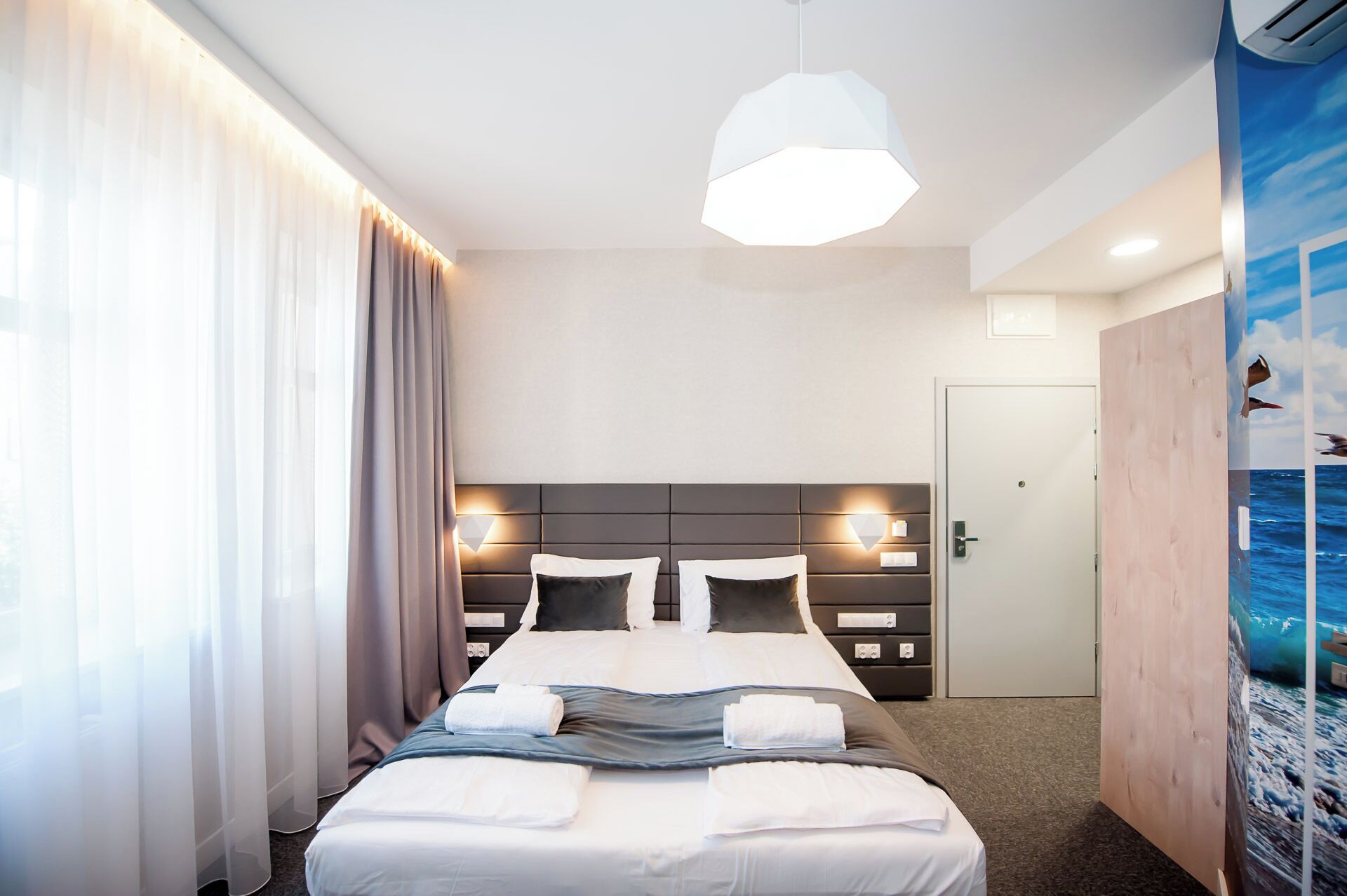 Une chambre d’hôtel dotée d’un grand lit, d’une tête de lit avec éclairage intégré, d’une vue sur l’océan par une fenêtre et d’une porte donnant accès à la salle de bain.
