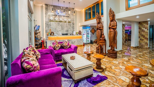 Un vestíbulo amplio con una mesa de recepción, sofás morados, una mesa de centro, estatuas decorativas y una pared de mármol con relojes.