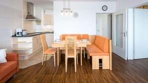 Una cucina moderna con mobili bianchi, forno integrato e macchina per il caffè. C'è un'area pranzo con tavolo in legno, cuscini arancioni e sedie.