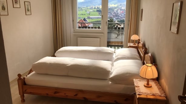 A double bed with a wooden frame, a bedside table with a lamp, and a window with a view of a town and mountains. - Disentiserhof Hönig by Interhome (Disentis)