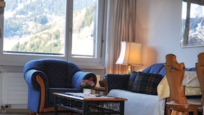 A living room with a view of mountains through large windows, a blue armchair, a white sofa, a wooden coffee table, and a lamp. - Disentiserhof Hönig by Interhome (Disentis)