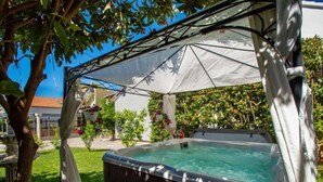 Een jacuzzi onder een witte luifel, omringd door groen en een terras van natuursteen.