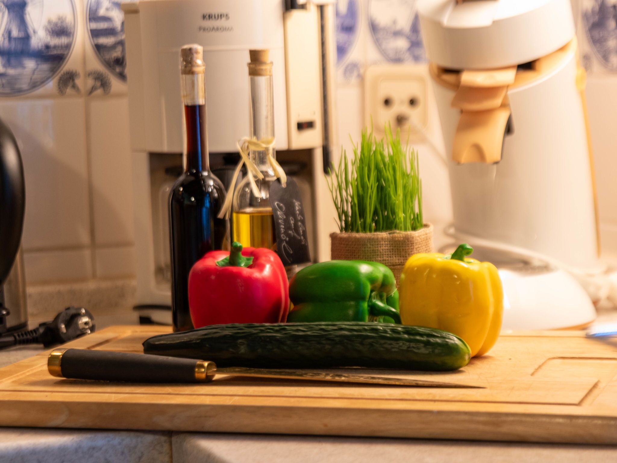 Uma bancada de cozinha com uma faca, um pimentão vermelho, um pimentão verde, um pimentão amarelo, um pepino e duas garrafas de azeite.