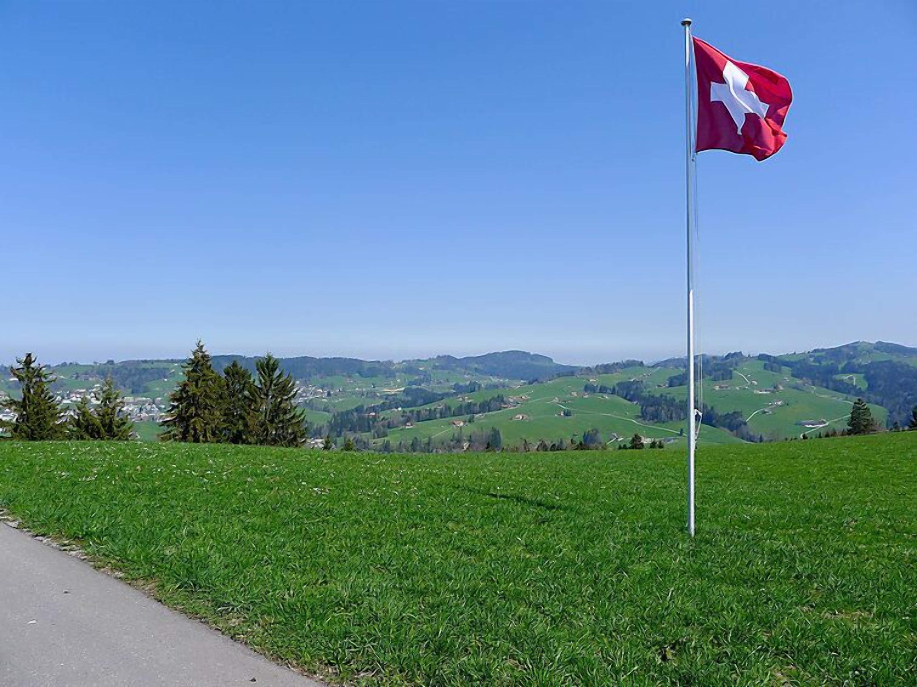 Una bandera en un asta, en un área con césped, con vistas a colinas ondulantes y un pueblo distante.