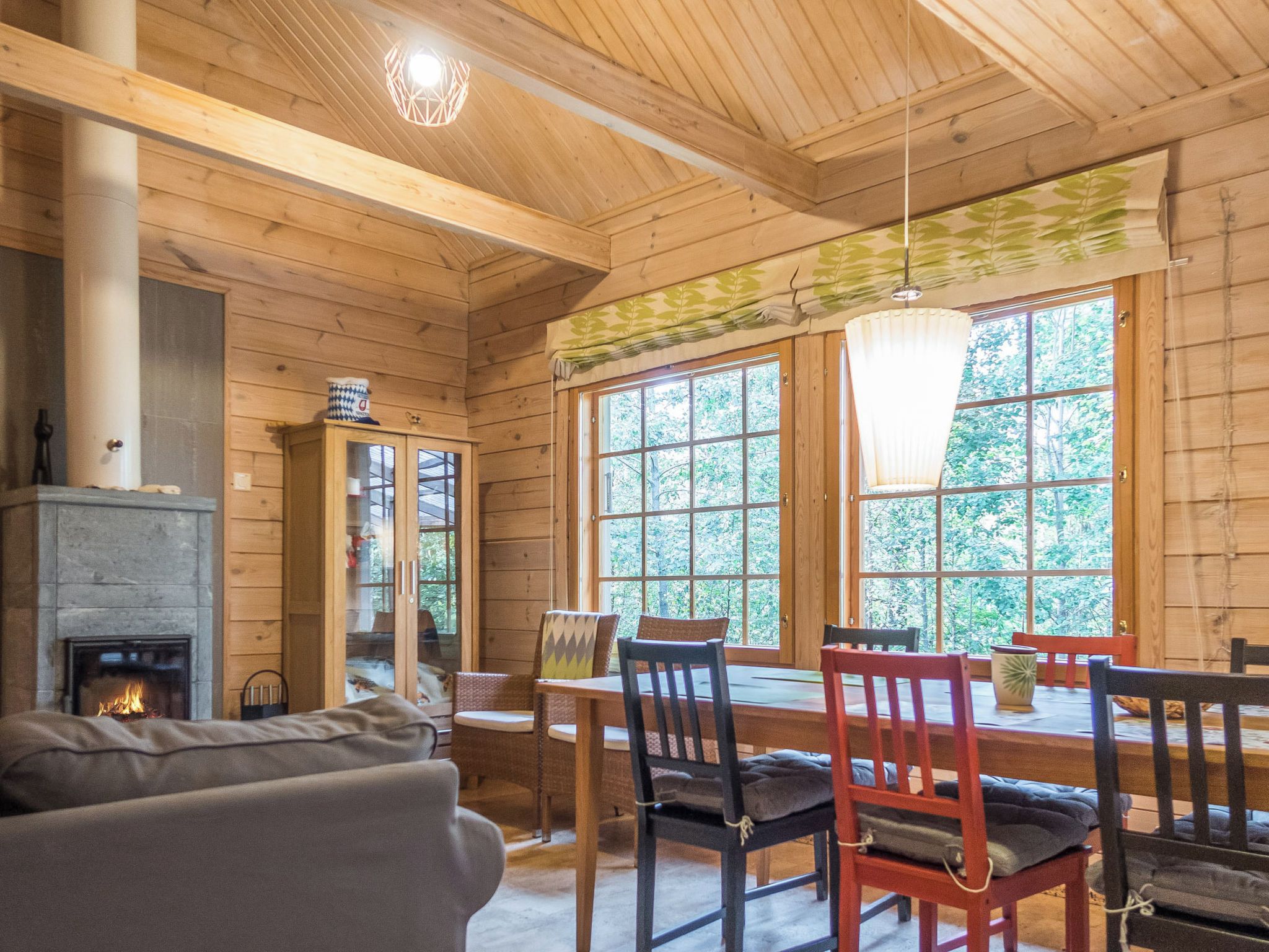 A cozy cabin interior with a fireplace, wooden walls, a dining area with chairs, and large windows offering a view of the outdoors.