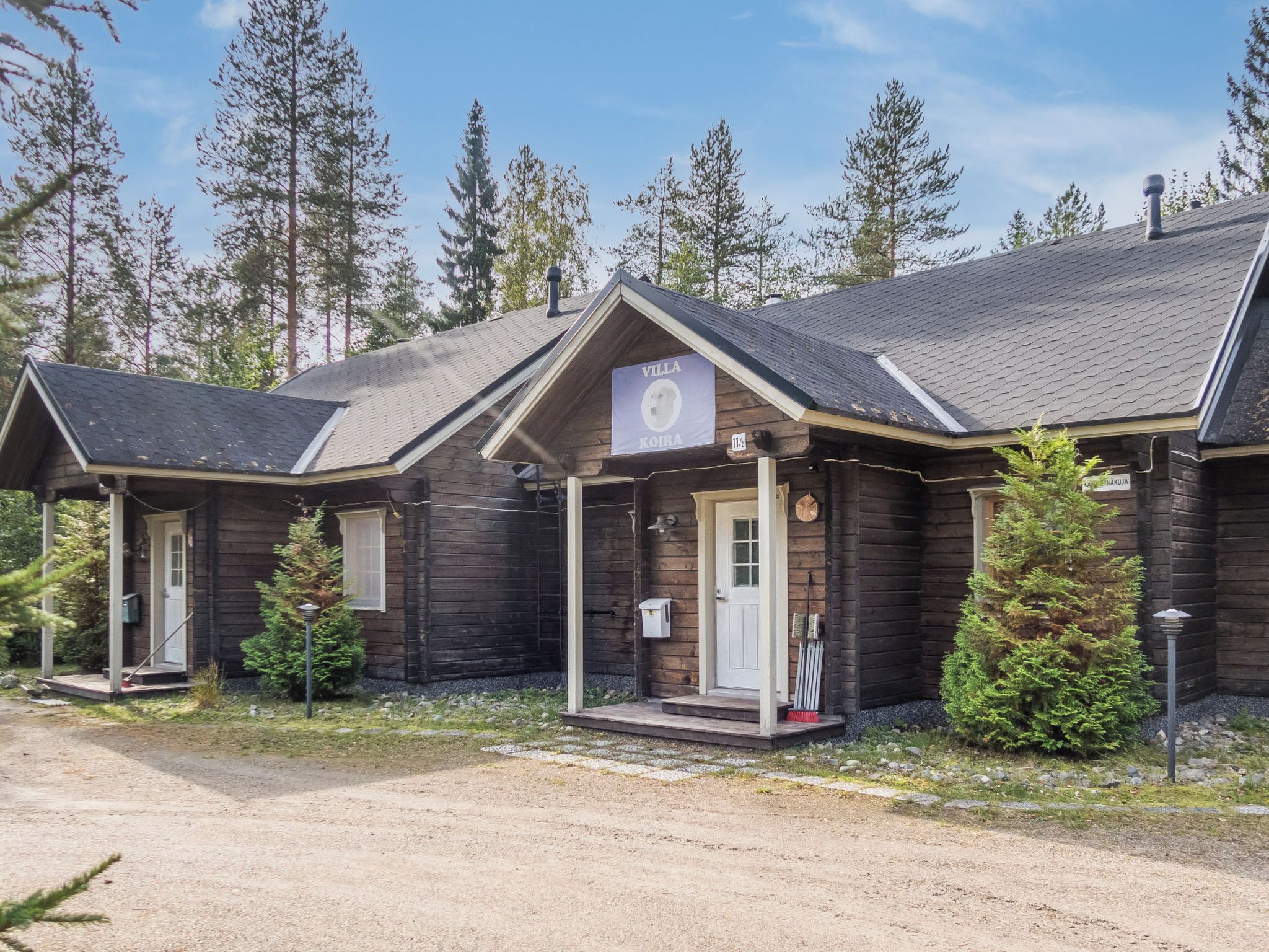Rivissä on puisia mökkejä, ja yhdessä niistä on kyltti, jossa lukee ”Villa Koira”, ja sen alla on numero ”11/2”, joka osoittaa mökin numeron.