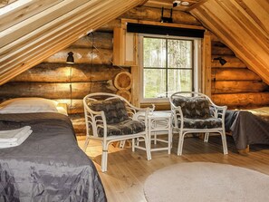 Interior de una cabaña de troncos con un dormitorio en el altillo, dos sillas blancas de mimbre, un ventanal con vistas a árboles y techo de madera.