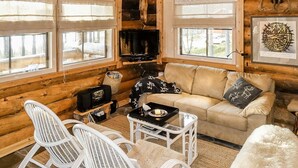 L’intérieur d’un chalet confortable, avec un canapé beige, des fauteuils en osier blancs, une petite table sur laquelle est posée une bougie et un téléviseur.