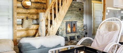 Ein gemütliches Blockhaus-Interieur mit Kamin aus Stein, einer Holztreppe und einem weißen Sessel mit Kissen.