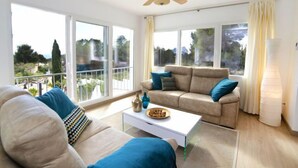Ein geräumiges Wohnzimmer mit einem beigefarbenen Sofa, einem weißen Couchtisch und einem Deckenventilator. Große Fenster bieten Blick ins Freie.