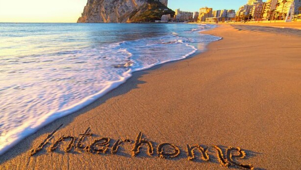 Ein Strand mit dem Schriftzug "interhome" im Sand, einem felsigen Felsen und Gebäuden im Hintergrund.