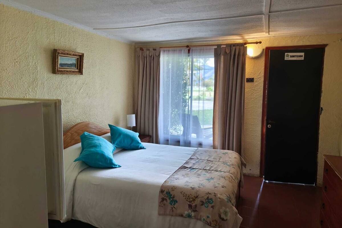 Ett hotellrum med en säng, ett nattduksbord, en lampa, ett fönster med gardiner och en dörr med texten ”DESINFICERAD”.