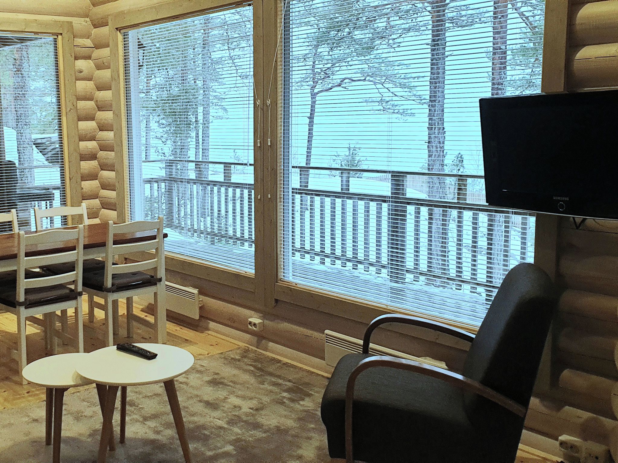 A log cabin interior with a dining area, a television, and large windows with a view of trees and buildings.