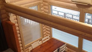 L’intérieur d’un chalet confortable, avec des murs en rondins de bois, une table et des chaises en bois, une cheminée en briques et de grandes fenêtres équipées de stores.
