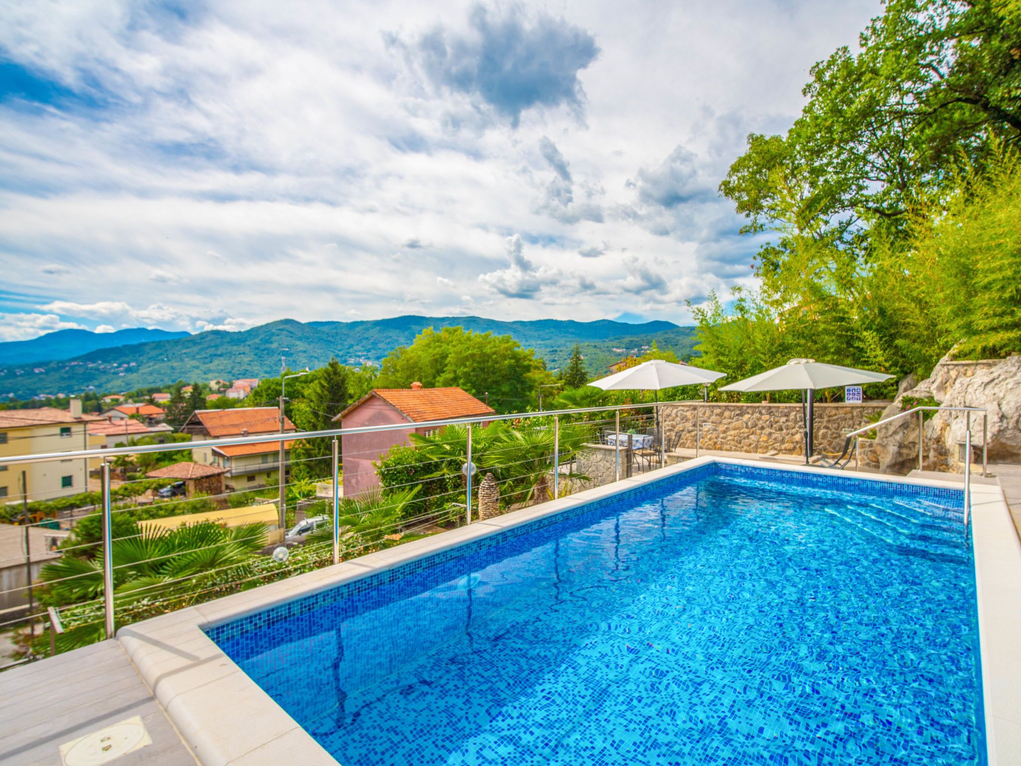 A rectangular swimming pool with a tiled edge, surrounded by a stone wall and metal railings. There are two umbrellas providing shade. The pool is set against a backdrop of lush greenery and distant mountains.