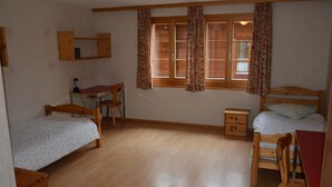 A room with two single beds, a wooden desk with a chair, a small bedside table, and a window with curtains. - Anne (Tiefparterre) by Interhome (Schönried)