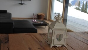 A wooden table with a white lantern with a heart-shaped cutout, a black sofa, a flat-screen TV on the wall, and a view of a snowy mountain through a large window.