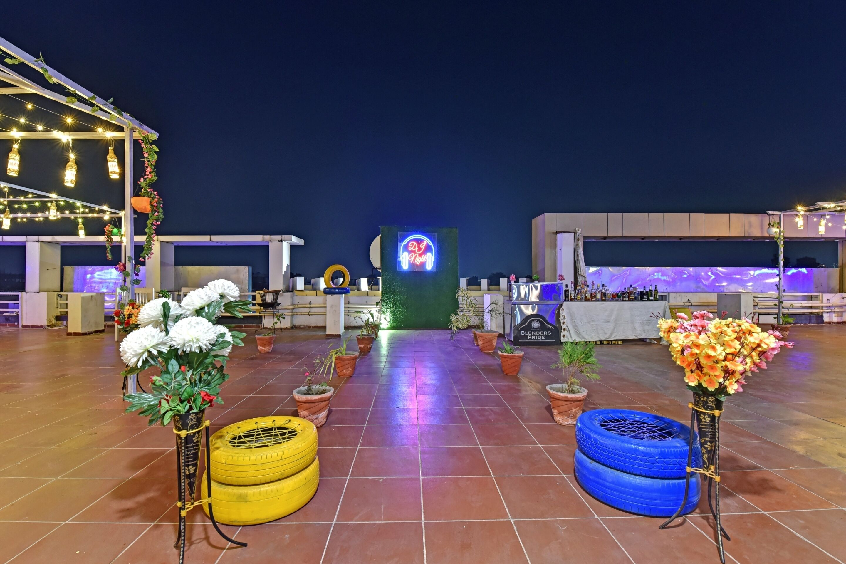 A rooftop terrace with potted plants, a neon sign, and a stage area.