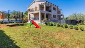 Un edificio de dos pisos con un balcón en el nivel superior, un patio de juegos con tobogán y un área cercada.
