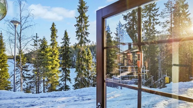 Vista a través de una ventana que muestra un paisaje nevado con altos árboles de hoja perenne y un edificio al fondo.