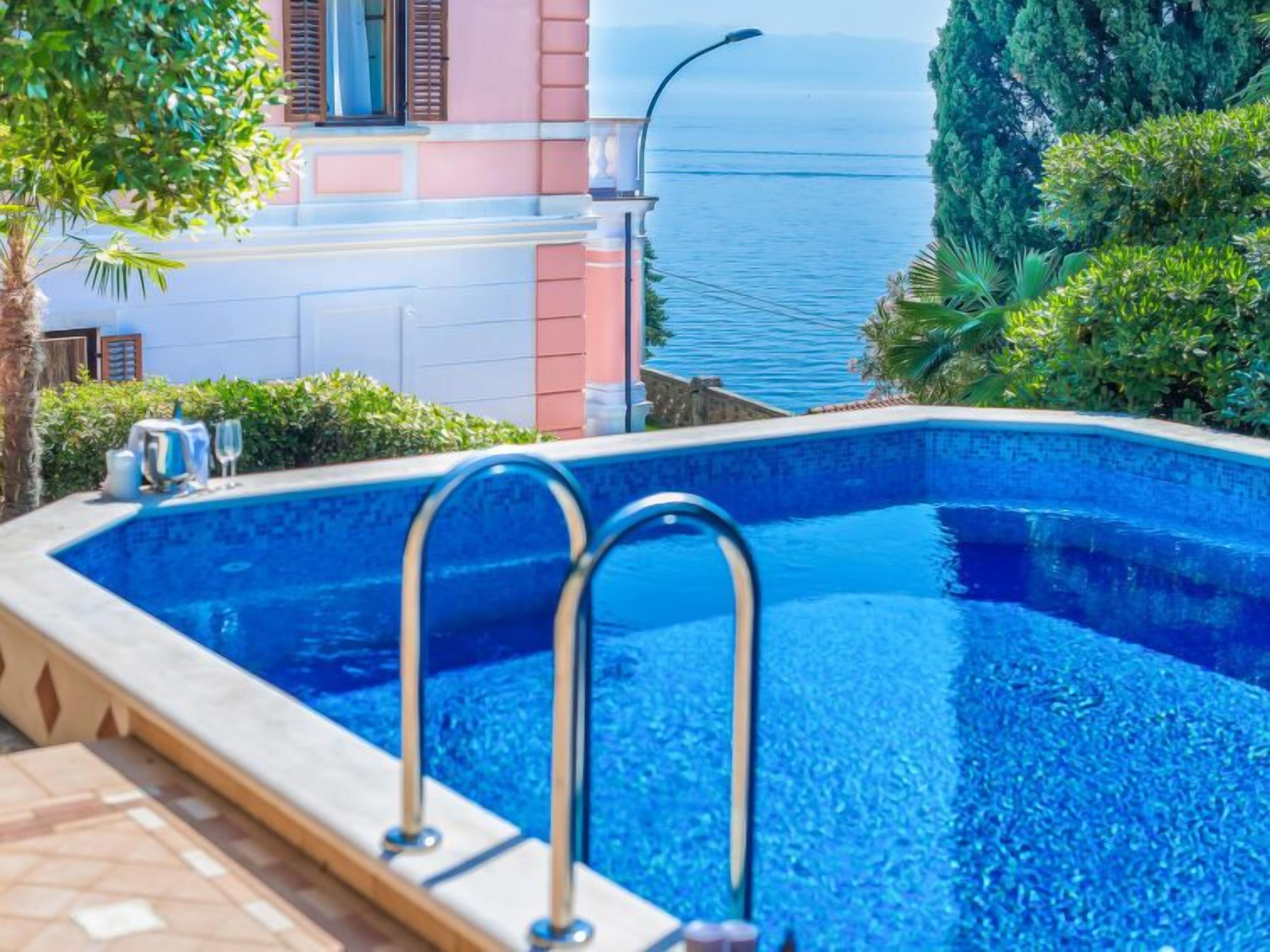Piscina com vista para o mar, um edifício com paredes rosa e brancas e uma palmeira.