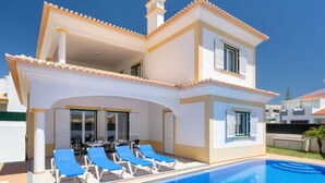 A two-story house with a swimming pool, blue lounge chairs, and a covered patio area.