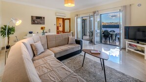 A modern living room with a sofa, a coffee table, a TV, and a sliding glass door leading to an outdoor area with a table and chairs.