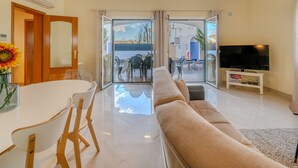 A modern living room with a sofa, a dining table with chairs, a TV on a stand, and a sliding glass door leading to an outdoor patio area.
