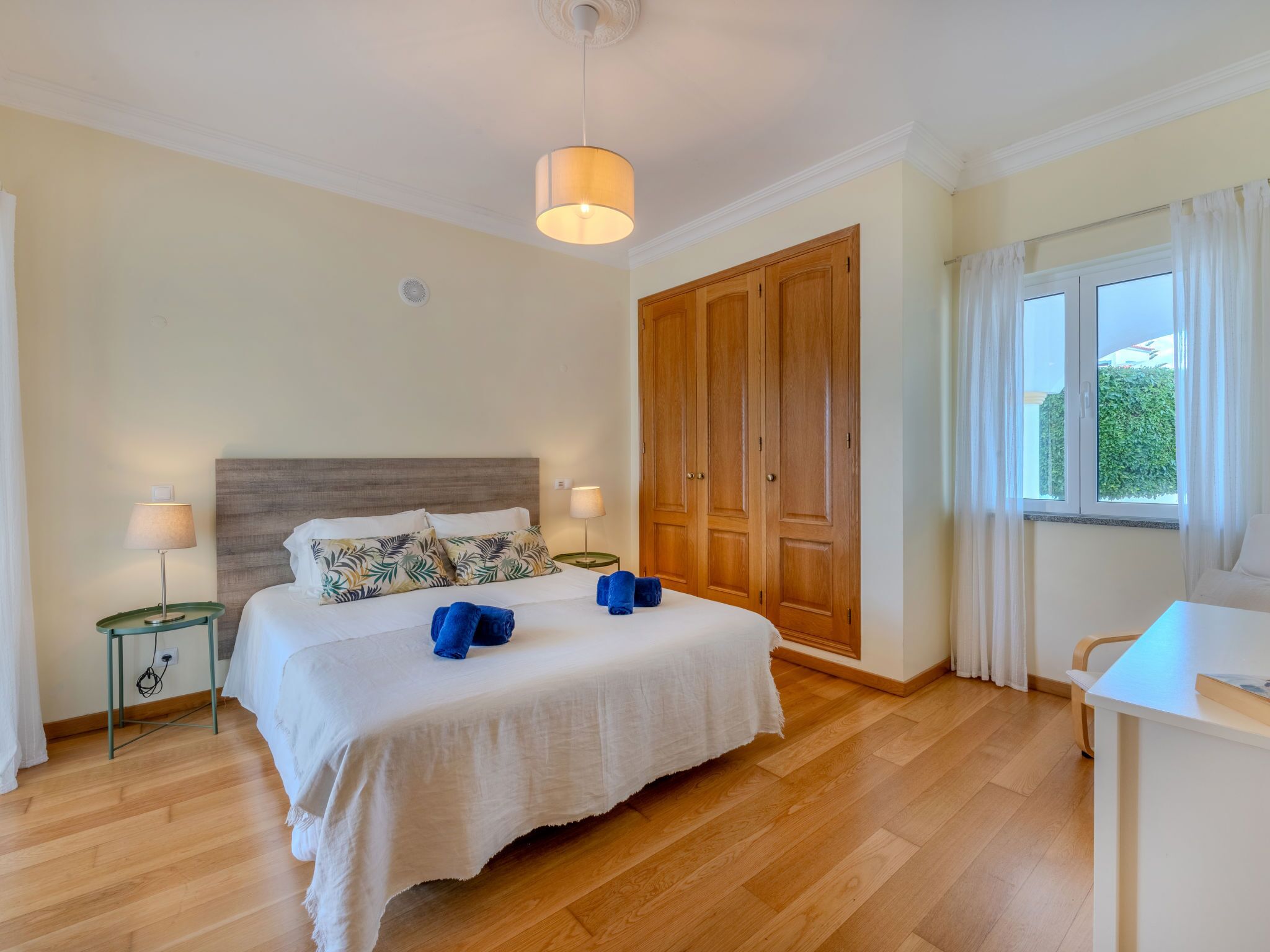 A hotel room with a wooden floor, a bed with white bedding, two bedside lamps, a wooden wardrobe, and a window with curtains.