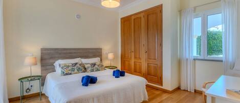 A hotel room with a wooden floor, a bed with white bedding, two bedside lamps, a wooden wardrobe, and a window with curtains.