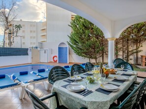 An outdoor dining area with a pool, blue lounge chairs, and a table set for two with plates, glasses, and a fruit bowl. - Colibri by Interhome (Gale)