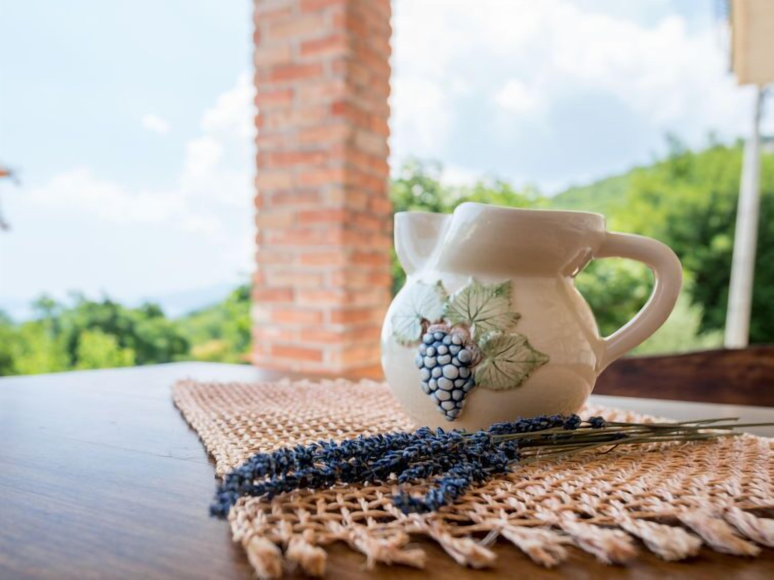 Una jarra de cerámica blanca con diseños de uva sobre un posavasos tejido, colocada sobre una mesa de madera con vistas a follaje y un muro de ladrillo al fondo.